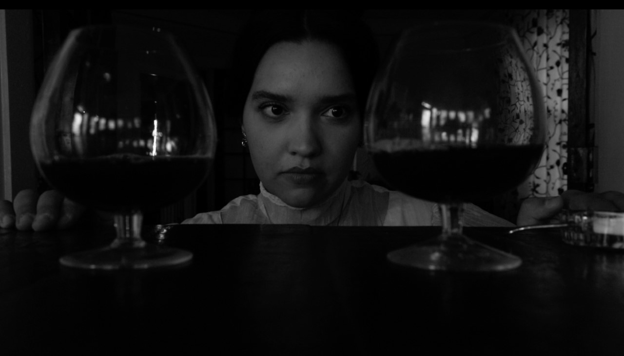 Black and white, a woman's face framed by wine glasses on either side.