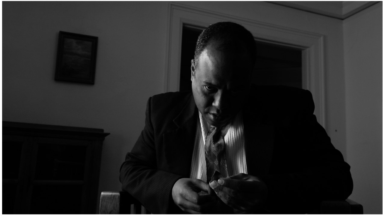 Black and white, a man in a suit contemplates a dagger held in his hand