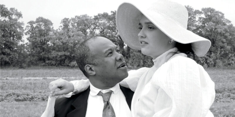 Black and white, a woman in a large sunhat sits on the lap of a man in a suit and tie, a copse of trees is visible in the backgroud.