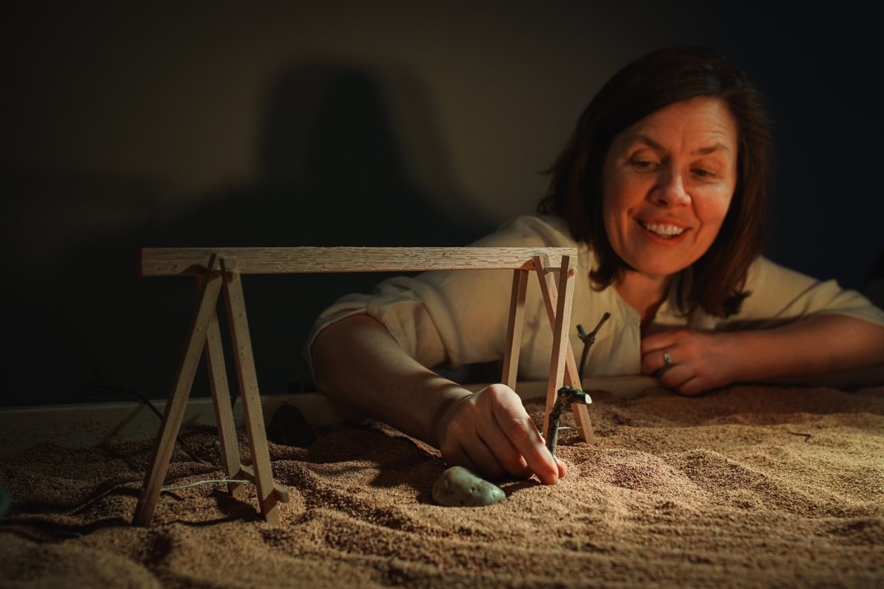 A woman builds a small wooden structure atop sand.