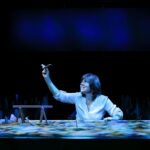 Actor at a chest-height table, top covered in sand where she places sticks and stones to illustrate her story, illuminated in blue.