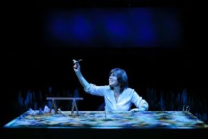 Actor at a chest-height table, top covered in sand where she places sticks and stones to illustrate her story, illuminated in blue.