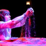 Actor at a chest-height table, top covered in sand, she lifts the sand in the air, letting it fall through her fingers, illuminated in purple.