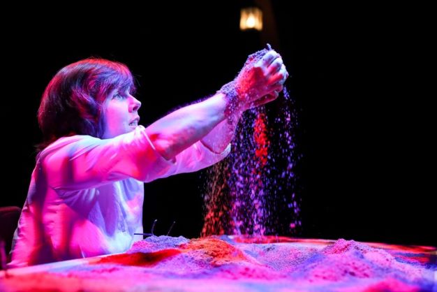 Actor at a chest-height table, top covered in sand, she lifts the sand in the air, letting it fall through her fingers, illuminated in purple.
