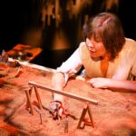 Actor at a chest-height table, top covered in sand where she places sticks and stones to illustrate her story, illuminated in a warm orange.