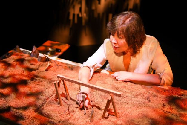 Actor at a chest-height table, top covered in sand where she places sticks and stones to illustrate her story, illuminated in a warm orange.