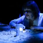 Actor at a chest-height table, top covered in sand where she places sticks and stones to illustrate her story, illuminated in blue.