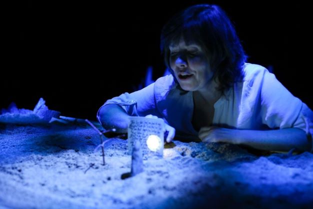 Actor at a chest-height table, top covered in sand where she places sticks and stones to illustrate her story, illuminated in blue.
