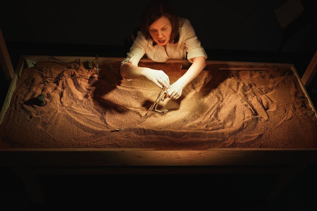 A woman seated at a table, drawing in sand