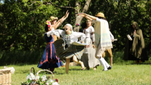 Revellers in colorful dress dance in front of a large tree and a hooded figure playing tamborine in the background.